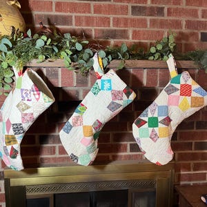 May include: Three handmade Christmas stockings with a patchwork design. Each stocking is white with colorful squares and diamond shapes. They hang above a fireplace with greenery and a brick background.
