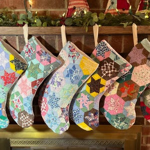 May include: Seven Christmas stockings hanging in a row, each crafted from a patchwork of multi-colored fabric hexagons. The stockings feature various floral, plaid, and geometric patterns in shades of red, blue, yellow, green, and pink.
