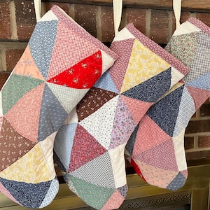 May include: Three Christmas stockings constructed from a patchwork of various fabric triangles in diverse colors and patterns, including red, blue, yellow, and pink floral and geometric prints, each featuring a white fabric cuff and hanging loop.