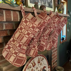 May include: A row of six Christmas stockings with a red and cream-colored geometric pattern. The stockings are hanging from a wooden mantel above a brick fireplace. The stockings have a traditional holiday design.