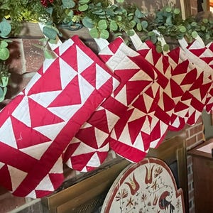 May include: A collection of red and white quilted Christmas stockings hang from a brick fireplace. The stockings feature a geometric star pattern. Greenery with red berries decorates the mantel above the stockings, creating a festive holiday display.