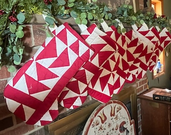 May include: A collection of red and white quilted Christmas stockings hang from a brick fireplace. The stockings feature a geometric star pattern. Greenery with red berries decorates the mantel above the stockings, creating a festive holiday display.