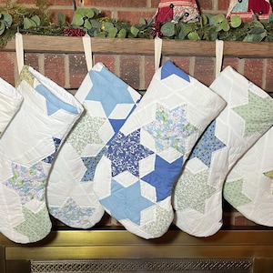 May include: Six quilted Christmas stockings hanging in a row, each featuring a white background with various blue, green, and yellow star-shaped fabric patches. The stockings are constructed with a patchwork design and have white fabric loops.