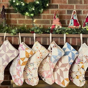 May include: Six quilted Christmas stockings hanging in a row, each featuring a unique patchwork design with various fabric patterns in shades of pink, blue, yellow, and orange, all suspended by white fabric loops from a wooden mantelpiece.