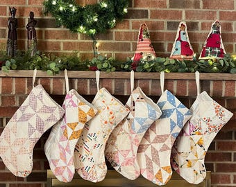 May include: Six quilted Christmas stockings hanging in a row, each featuring a unique patchwork design with various fabric patterns in shades of pink, blue, yellow, and orange, all suspended by white fabric loops from a wooden mantelpiece.