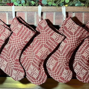 May include: Seven identical Christmas stockings hanging in a row, each featuring a deep red fabric base with a contrasting white woven geometric pattern. The stockings are suspended by simple white fabric loops from a wooden mantelpiece.