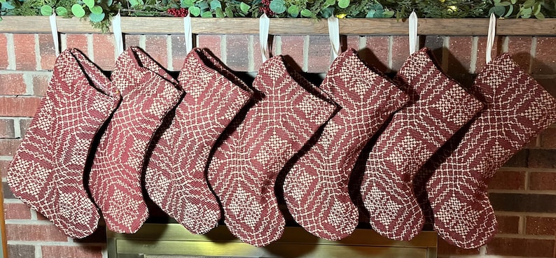 May include: Seven identical Christmas stockings hanging in a row, each featuring a deep red fabric base with a contrasting white woven geometric pattern. The stockings are suspended by simple white fabric loops from a wooden mantelpiece.