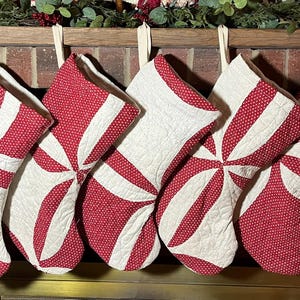 May include: Seven quilted Christmas stockings hanging in a row, each featuring a repeating pinwheel pattern. The stockings are constructed from alternating panels of solid off-white fabric and red fabric with a small white polka dot pattern.