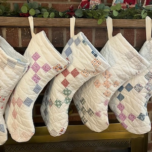 May include: Five quilted Christmas stockings hanging in a row. Each stocking is primarily white with a diamond-shaped patchwork pattern featuring various floral and checkered prints in shades of blue, red, purple, green, and orange.
