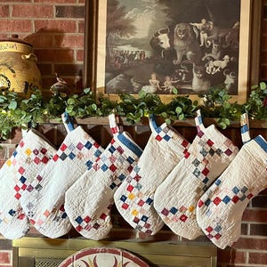 May include: Seven quilted Christmas stockings with a patchwork design of red, blue, and yellow squares on a white background. Each stocking has a blue and white striped cuff and is hanging from a mantelpiece.