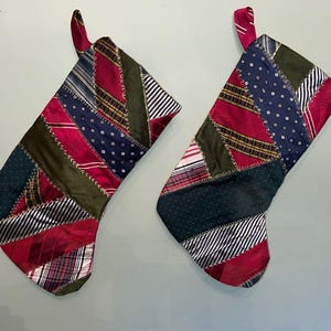 May include: Two holiday stockings constructed from a patchwork of various silk neckties in shades of deep red, navy blue, olive green, and white. The fabric pieces are joined with visible decorative stitching, and each stocking features a fabric loop.