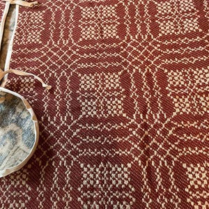 May include: A woven textile featuring a repeating geometric pattern in deep rust red and cream white. The fabric has a textured, grid-like appearance with intricate interlocking shapes. Attached to the side are two white fabric ties or straps.