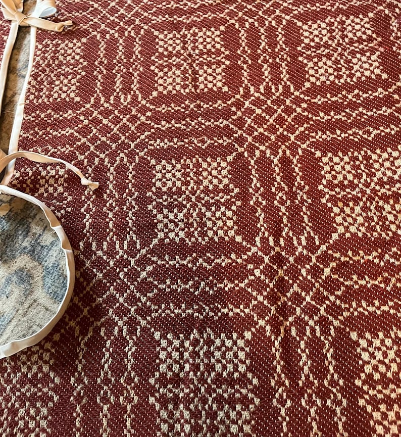 May include: A woven textile featuring a repeating geometric pattern in deep rust red and cream white. The fabric has a textured, grid-like appearance with intricate interlocking shapes. Attached to the side are two white fabric ties or straps.