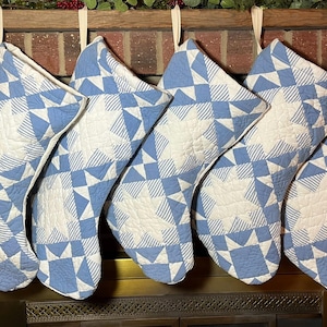 May include: Six quilted Christmas stockings featuring a blue and white geometric star pattern. Each stocking is constructed with a combination of solid blue fabric and blue-and-white striped fabric, creating a repeating star design throughout.