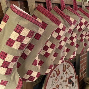 May include: A row of seven quilted Christmas stockings hanging in a line. Each stocking features a patchwork design with alternating squares of dark red plaid fabric and cream-colored fabric, accented by solid olive green fabric borders and cuffs.