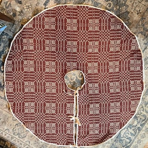 May include: A circular tree skirt featuring a deep red and white geometric woven pattern. The fabric has a white piped edge, a central circular cutout, and a straight slit opening secured with three sets of white fabric ties for closure.