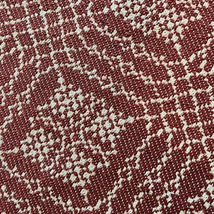 May include: A close-up view of a woven textile featuring a repeating pattern of interconnected, irregular shapes in a deep brick red color contrasted against a light beige background, creating a textured appearance with a visible grid-like weave.