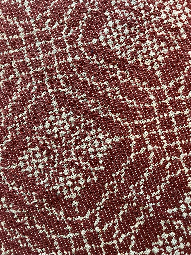 May include: A close-up view of a woven textile featuring a repeating pattern of interconnected, irregular shapes in a deep brick red color contrasted against a light beige background, creating a textured appearance with a visible grid-like weave.