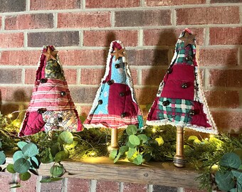 May include: Three handmade Christmas tree decorations. Each tree is made of patchwork fabric in red, green, and blue patterns, adorned with wooden star toppers and small bells. They stand on wooden bases, arranged on a rustic wooden shelf with greenery.