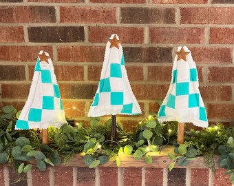 May include: Three decorative Christmas trees with white and teal checkered patterns. Each tree has a wooden trunk and a star on top. The trees are arranged on a wooden surface with greenery and fairy lights in front of a brick wall.