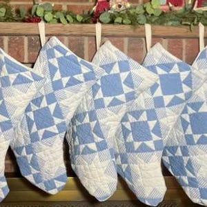 May include: A row of seven quilted Christmas stockings featuring a light blue and white geometric star pattern. Each stocking is constructed from fabric with a stitched texture and includes a fabric loop at the top for hanging purposes.