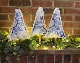 May include: Three decorative Christmas tree ornaments. Each tree is white with blue floral embroidery and a scalloped edge. The trees are mounted on wooden dowels and sit on a shelf with greenery and lights.