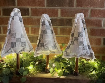 May include: Three decorative Christmas tree ornaments. Each tree has a white and gray patchwork design, a wooden stem, and a ruffled edge. The trees are arranged on a shelf with greenery and fairy lights, against a brick wall.