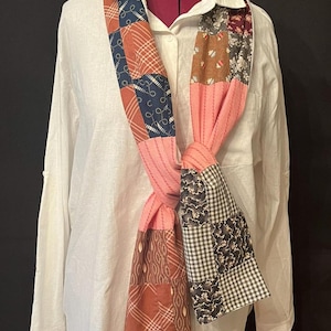 May include: A long patchwork scarf featuring a variety of fabric squares in shades of pink, brown, navy blue, and white. The scarf displays diverse patterns including plaid, gingham, floral, and geometric prints, joined by visible stitching.