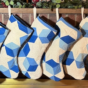 May include: Seven quilted Christmas stockings hanging in a row, each featuring a geometric pattern of interlocking cubes in shades of light blue, medium blue, and dark blue against a cream-colored background, with a solid blue fabric cuff.
