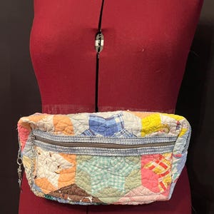 May include: A fanny pack constructed from multicolored quilted fabric featuring a star-patterned patchwork design. The bag includes a horizontal zippered closure across the front with a metal zipper pull and a light blue denim-textured fabric trim.