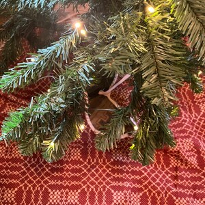 May include: Artificial green pine tree branches with small warm white LED string lights woven throughout the needles. The branches are positioned over a red and white woven fabric featuring a repeating geometric pattern across the surface.