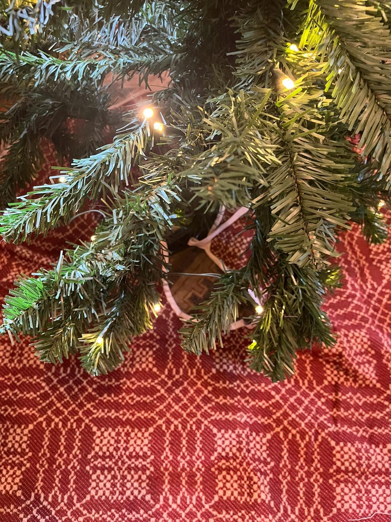 May include: Artificial green pine tree branches with small warm white LED string lights woven throughout the needles. The branches are positioned over a red and white woven fabric featuring a repeating geometric pattern across the surface.