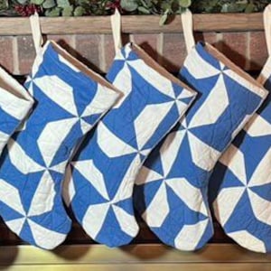 May include: Seven quilted Christmas stockings hanging in a row, each featuring a blue and white pinwheel pattern. The stockings are constructed with a stitched fabric design and include a light-colored fabric loop at the top for hanging purposes.