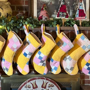 May include: A row of eight quilted Christmas stockings. Each stocking has a yellow background with patchwork squares of pink, blue, and white. They hang from a fireplace mantel, creating a festive holiday display.