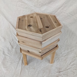 May include: A wooden compost bin with three tiers and a lid. The bin is made of natural wood and has a hexagonal shape. It is sitting on four legs.