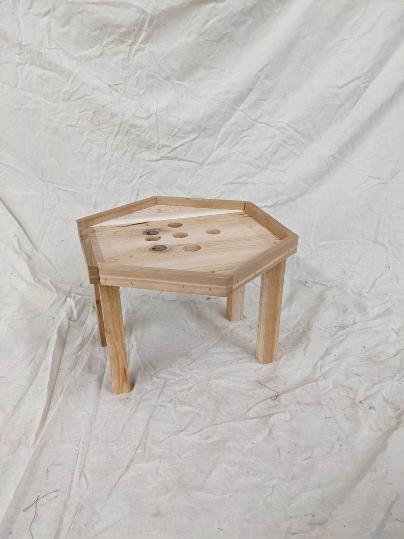 May include: A hexagonal wooden table with a light wood finish and six legs. The table top has a series of holes drilled into it.