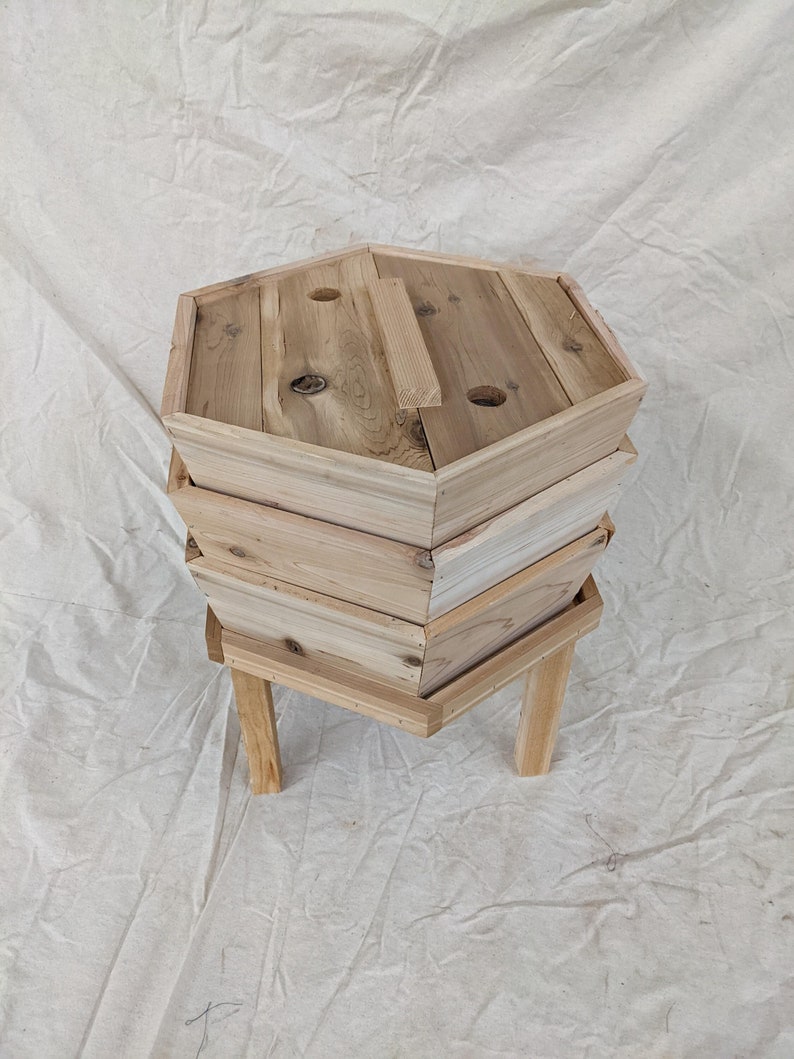 May include: A wooden compost bin with three tiers and a lid. The bin is made of natural wood and has a hexagonal shape. It is sitting on four legs.