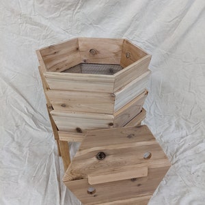 May include: A wooden hexagonal compost bin with three tiers and a removable lid. The bin is made of natural wood and has a wire mesh screen in the bottom tier.