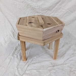 May include: A hexagonal wooden planter with a removable top and four legs. The planter is made of natural wood and has a rustic appearance.