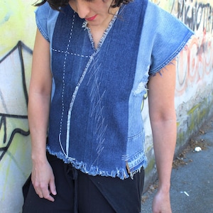 Può includere: Un top senza maniche in denim con scollo a V e bordi grezzi. Il top è realizzato con varie tonalità di denim blu e presenta dettagli di cucitura bianchi. La modella indossa pantaloni neri.