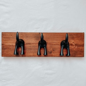May include: Three black metal dog-shaped hooks mounted on a dark brown wooden board.