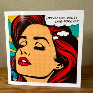 May include: Pop art style print featuring a woman with red hair and red lips. The woman is looking up with closed eyes. The print has the text "DREAM LIKE YOU'LL LIVE FOREVER" in a speech bubble. The background is yellow, blue, and black.