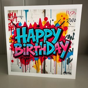 May include: A white greeting card featuring a graffiti-style design. The words "HAPPY BIRTHDAY" are in bold blue and pink letters, outlined in black. The background includes colourful paint drips and graffiti art.