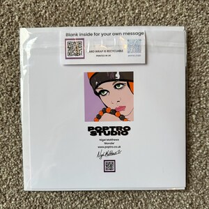 May include: A greeting card featuring a pop art illustration of a woman with green eyes, wearing a black and orange hat and a beaded necklace. The card has the text "Blank inside for your own message" and "ARD WRAP IS RECYCLABLE PRINTED IN UK".