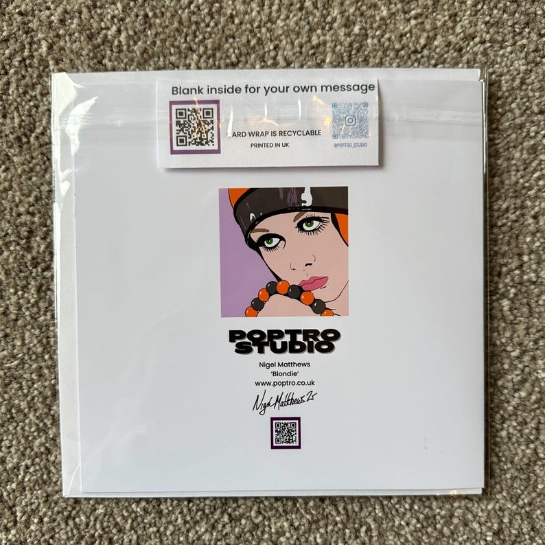 May include: A greeting card featuring a pop art illustration of a woman with green eyes, wearing a black and orange hat and a beaded necklace. The card has the text "Blank inside for your own message" and "ARD WRAP IS RECYCLABLE PRINTED IN UK".