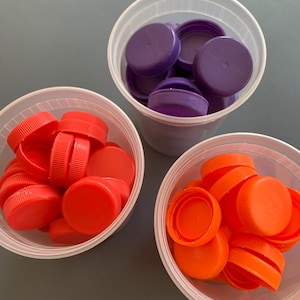May include: Three clear plastic containers filled with colorful plastic bottle caps. One container has purple caps, one has orange caps, and one has red caps.