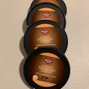 Puede incluir: Cuatro recipientes de plástico negro de queso parmesano rallado de Wellsley Farms. Cada recipiente tiene una etiqueta marrón con el logotipo de la empresa y el texto "Queso parmesano rallado" y "Envejecido naturalmente durante más de 10 meses". El recipiente inferior tiene una pegatina 100% "Envejecido naturalmente" y el texto "Peso neto 20 oz (1 lb 4 oz) (567 g)".