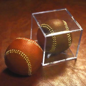 May include: Two baseballs, one in a clear acrylic cube, sit on a brown surface. The baseballs are brown with yellow stitching. The baseball in the cube is a darker shade of brown than the other baseball.