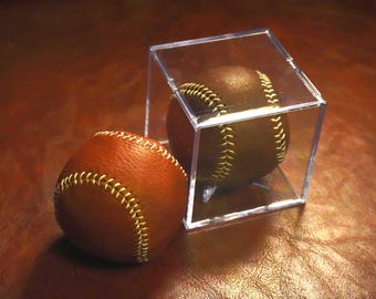 Pode incluir: Duas bolas de beisebol, uma em um cubo de acrílico transparente, estão em uma superfície marrom. As bolas de beisebol são marrons com costuras amarelas. A bola no cubo é de um tom marrom mais escuro.