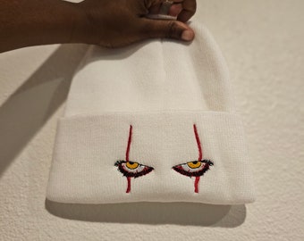 May include: White beanie with embroidered eyes featuring yellow irises, black eyelashes, and red vertical lines. The beanie is held up by a hand against a white background. The design is a stylized representation of a horror character.